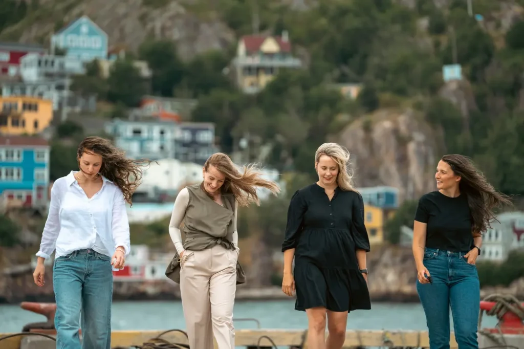 Kelia Cadigan, founder of The Harbour, walking with three mothers at St. John's Harbour. They are walking towards the camera, with their faces mostly looking down. The person on the right is looking at Kelia. Brightly coloured houses are visible in the background.