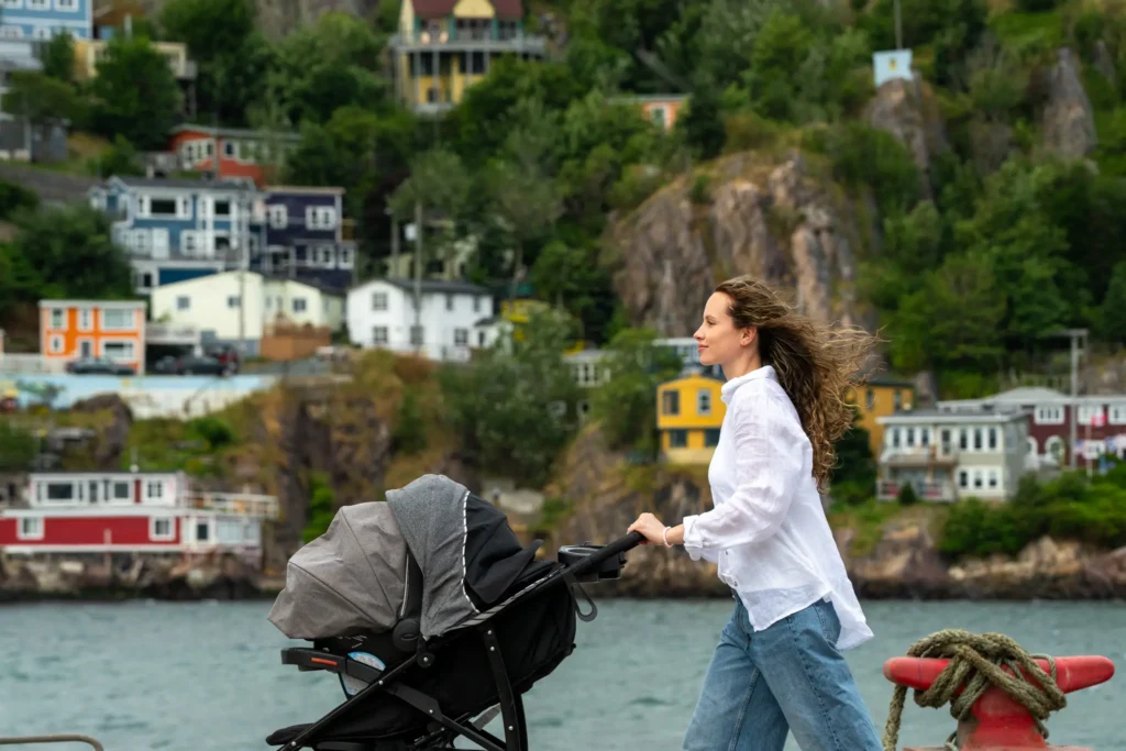 Kelia Cadigan, founder of The Harbour, walking and pushing a stroller at St. John's Harbour, with a row of brightly coloured houses in the background.