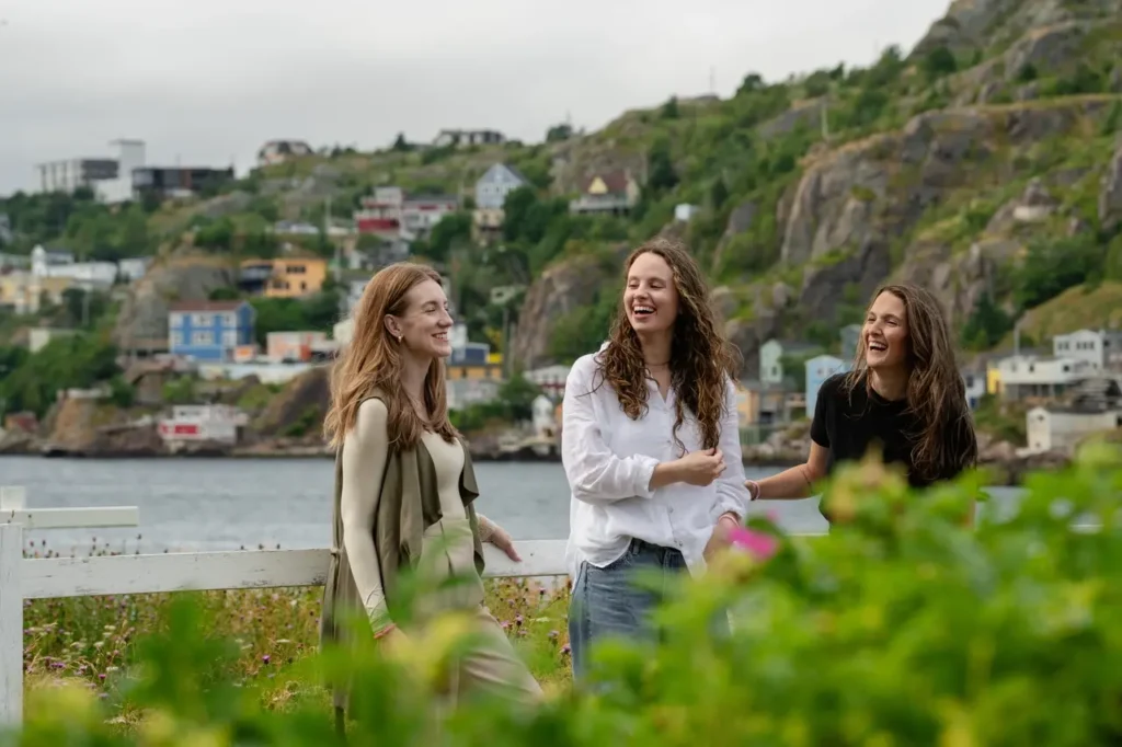 Three mothers from The Harbour community laughing and looking very happy at St. John’s Harbour, with green leaves and grass around and colourful houses visible in the background.