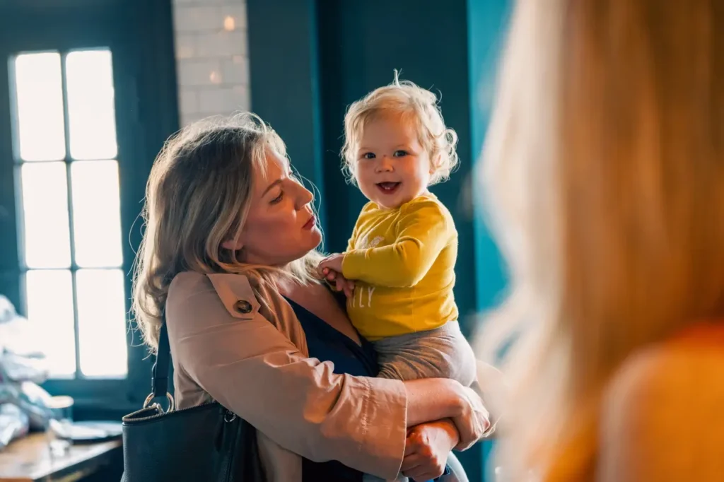 Mother holding her smiling child in her arms, with the child looking very happy. The photo is taken from behind another person standing nearby.