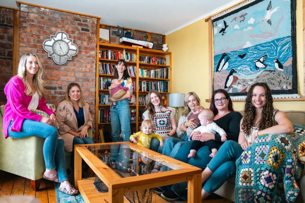 Seven mothers with their children, some in arms and some standing nearby, all looking at the camera and smiling. The room is bright with a knitted picture on the right wall and a shelf of books in the corner.