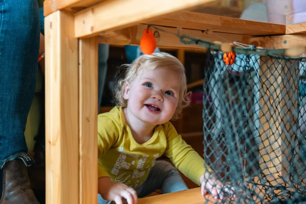 Curious child wearing a yellow long-sleeve shirt, smiling and sitting on the floor under a table, with a parent sitting nearby on the couch.