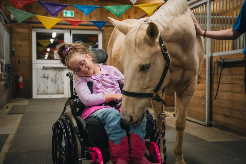 Child in a wheelchair interacting with a calm white horse in a beautifully decorated barn for the Hoedown in the Barn event.